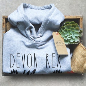 May include: A gray hoodie with the text "DEVON REX" in black, along with paw prints. The hoodie is inside a wooden crate with a succulent plant and a tag that reads "S&P Co. ORIGINAL GOODS MADE TO LAST."