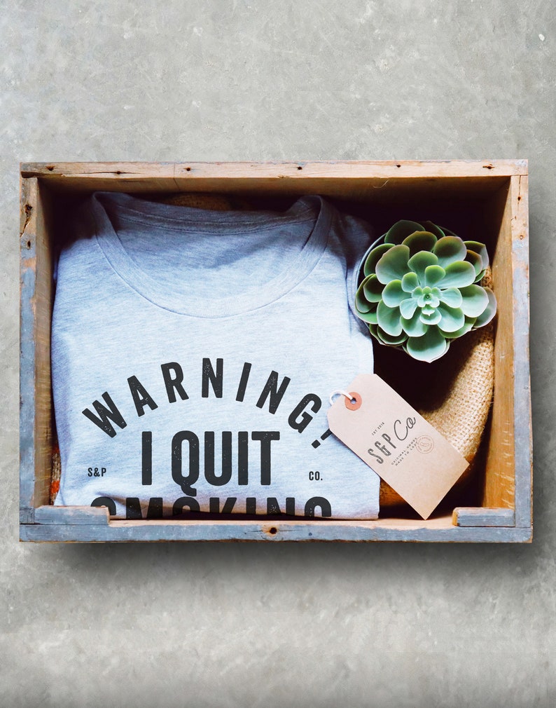 K&ouml;nnte beinhalten: Ein graues T-Shirt mit dem Text "WARNING I QUIT SMOKING" in schwarzen Buchstaben. Das Shirt befindet sich in einer Holzkiste mit einer kleinen Sukkulente und einem braunen Etikett mit dem Text "S&P Co."