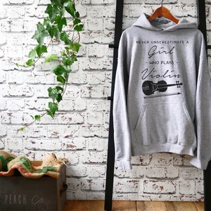 May include: Gray hooded sweatshirt with the text "Never underestimate a girl who plays violin" and a black silhouette of a violin.