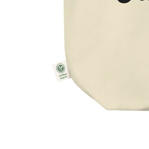 May include: A close-up of a beige tote bag with a green "certified organic" label. The label has a green leaf logo.