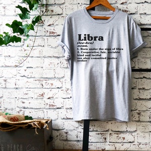 May include: Gray t-shirt with black text that reads "Libra (lee-bra) noun. 1. Born under the sign of libra 2. Cooperative, fair, sociable kind and tactful see also: committed justice seeker"