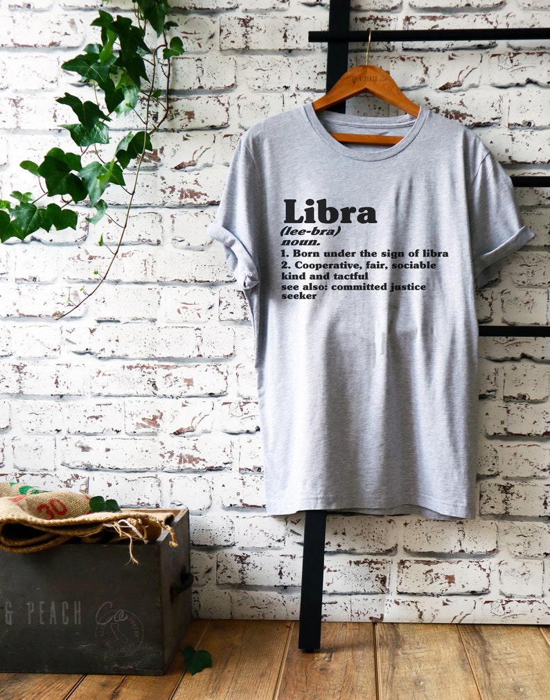 May include: Gray t-shirt with black text that reads "Libra (lee-bra) noun. 1. Born under the sign of libra 2. Cooperative, fair, sociable kind and tactful see also: committed justice seeker"