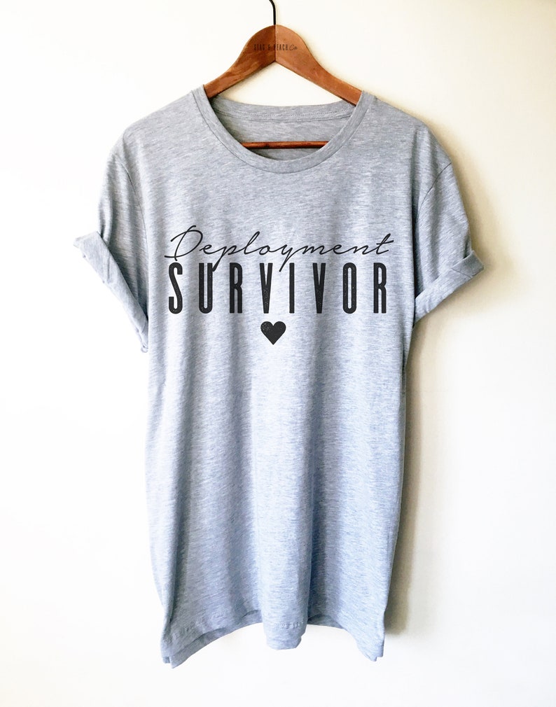Puede incluir: Una camiseta gris con el texto "Deployment Survivor" y un peque&ntilde;o coraz&oacute;n negro impreso en la parte delantera.