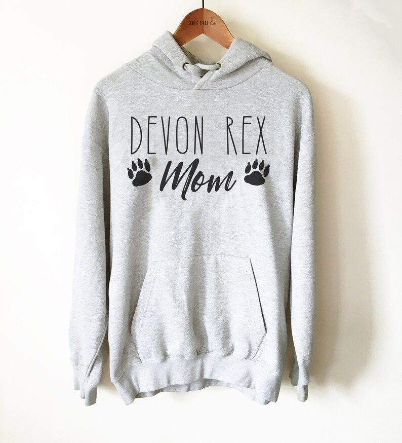 May include: A light gray hoodie with the text "DEVON REX Mom" in black, along with paw print graphics. The hoodie has a drawstring hood and a front pocket. The garment is hanging on a wooden hanger.