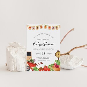 May include: A baby shower invitation with a watercolor illustration of a wheelbarrow full of vegetables and a chalkboard sign. The text reads "A baby is growing. Join us for a baby shower honoring Lucy Smith. June 21, 2-4pm. 1 Market Lane, Market Town, CA. Rsvp Lacey 888.888.8888. Registered at Baby World."
