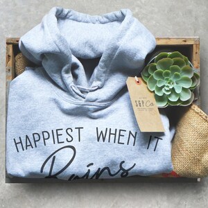 Puede incluir: Una sudadera con capucha gris con el texto "Happiest When It Rains" en letras negras. La sudadera est&aacute; en una caja de madera con una peque&ntilde;a planta suculenta y una etiqueta de papel marr&oacute;n.