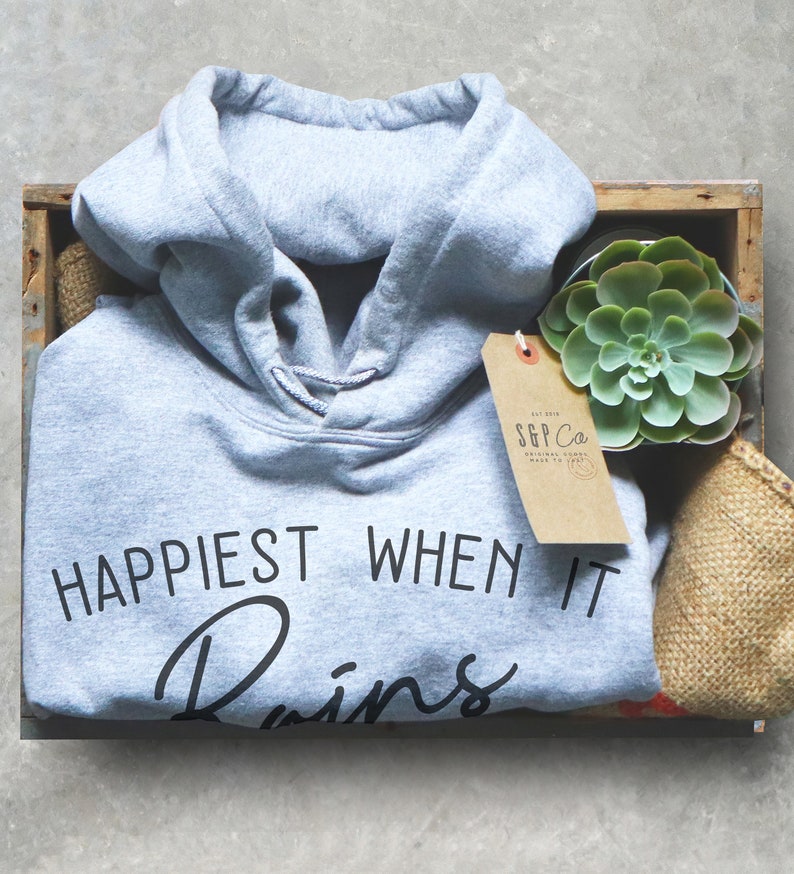 Puede incluir: Una sudadera con capucha gris con el texto "Happiest When It Rains" en letras negras. La sudadera est&aacute; en una caja de madera con una peque&ntilde;a planta suculenta y una etiqueta de papel marr&oacute;n.