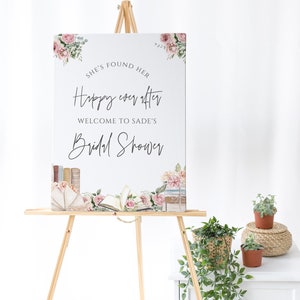 Op de afbeelding: Een wit bord met roze bloemenaccenten en de tekst "She's found her happy ever after. Welcome to Sade's Bridal Shower." Het bord staat op een ezel met een bloemstuk op de achtergrond.