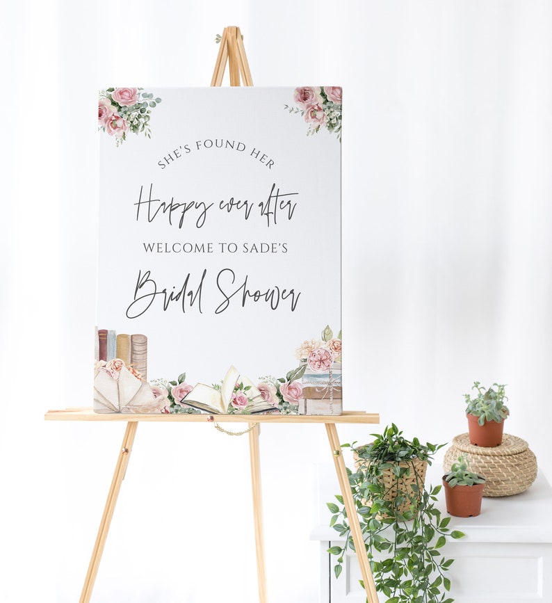 Op de afbeelding: Een wit bord met roze bloemenaccenten en de tekst "She's found her happy ever after. Welcome to Sade's Bridal Shower." Het bord staat op een ezel met een bloemstuk op de achtergrond.