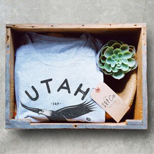 Op de afbeelding: Een grijs T-shirt met een zwarte adelaar afbeelding en de tekst "UTAH -S&P-." Het shirt zit in een houten kist met een kleine groene succulent plant en een bruin label met de tekst "S&P Co."