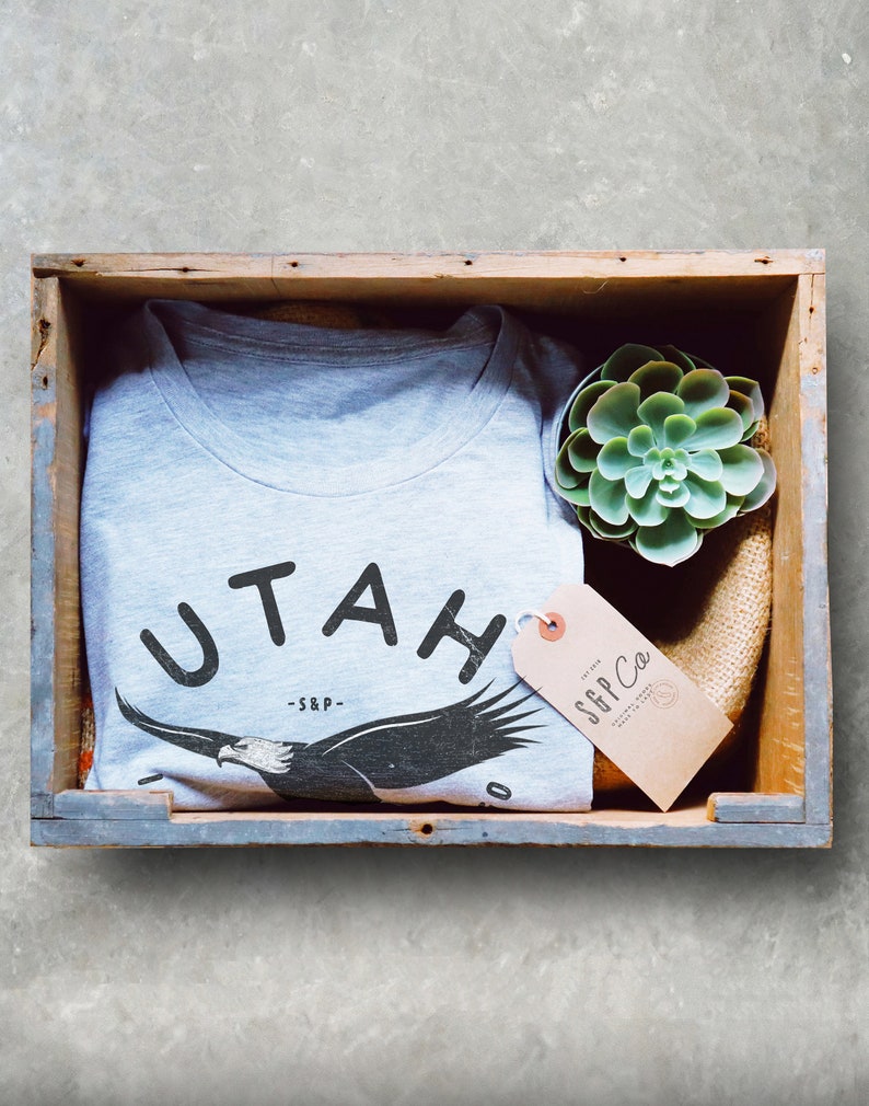 Op de afbeelding: Een grijs T-shirt met een zwarte adelaar afbeelding en de tekst "UTAH -S&P-." Het shirt zit in een houten kist met een kleine groene succulent plant en een bruin label met de tekst "S&P Co."
