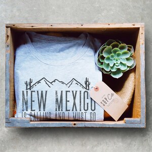 May include: A gray t-shirt with a black graphic design that says "New Mexico is calling and I must go". The graphic design features a mountain range and cacti. The t-shirt is in a wooden crate with a small succulent plant and a brown tag that says "SGP Co."