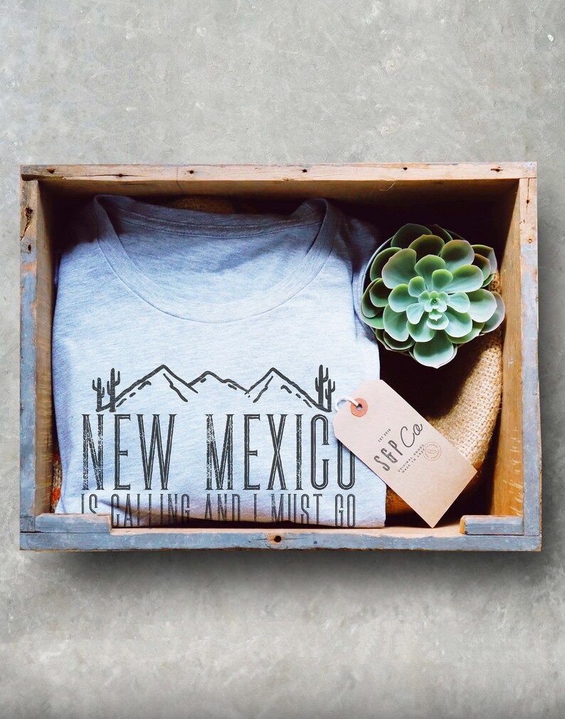 May include: A gray t-shirt with a black graphic design that says "New Mexico is calling and I must go". The graphic design features a mountain range and cacti. The t-shirt is in a wooden crate with a small succulent plant and a brown tag that says "SGP Co."