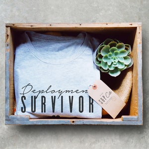Puede incluir: Una camiseta gris con el texto "Deployment Survivor" en letras negras. La camiseta est&aacute; en una caja de madera con una peque&ntilde;a planta suculenta y una etiqueta marr&oacute;n con el texto "SGP Co."