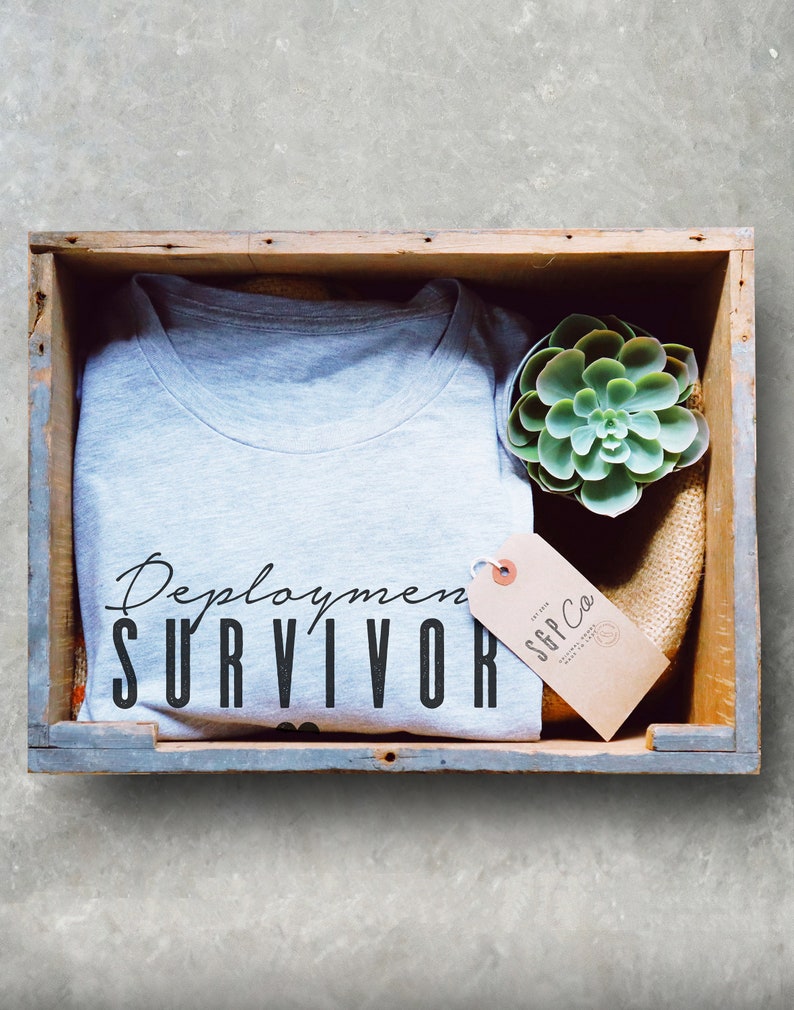 Puede incluir: Una camiseta gris con el texto "Deployment Survivor" en letras negras. La camiseta est&aacute; en una caja de madera con una peque&ntilde;a planta suculenta y una etiqueta marr&oacute;n con el texto "SGP Co."