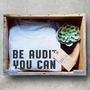 Może przedstawiać: Szary T-shirt z nadrukiem "BE AUDI YOU CAN". T-shirt znajduje się w drewnianej skrzyni z małą sukulentem i brązową etykietą z tekstem "SGP Co."