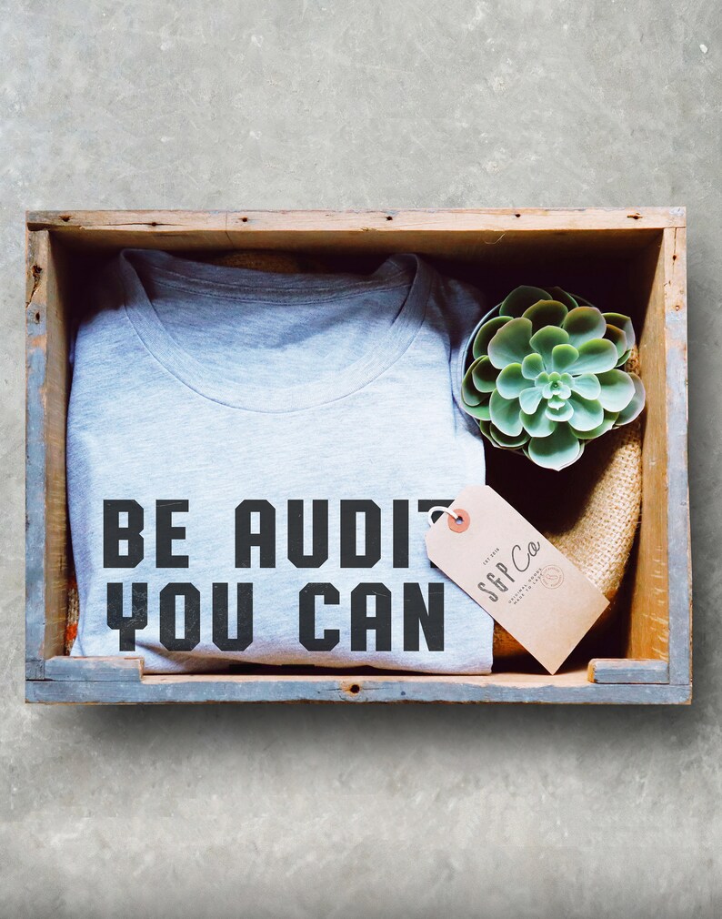 Może przedstawiać: Szary T-shirt z nadrukiem "BE AUDI YOU CAN". T-shirt znajduje się w drewnianej skrzyni z małą sukulentem i brązową etykietą z tekstem "SGP Co."