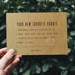 May include: A brown card with the text "YOUR NEW FAVORITE HOODIE" and a list of features: "ORIGINAL CRAFTED DESIGNS BY US", "MADE WITH SOFT AIR-JET SPUN YARN", "PRINTED WITH ECO-FRIENDLY INKS", "MADE TO LAST". The card also has a small round stamp with the text "STAG & PEACH Co."