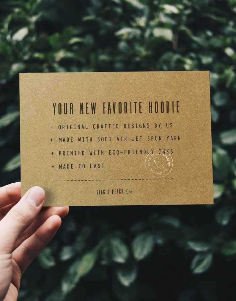 May include: A brown card with the text "YOUR NEW FAVORITE HOODIE" and a list of features: "ORIGINAL CRAFTED DESIGNS BY US", "MADE WITH SOFT AIR-JET SPUN YARN", "PRINTED WITH ECO-FRIENDLY INKS", "MADE TO LAST". The card also has a small round stamp with the text "STAG & PEACH Co."