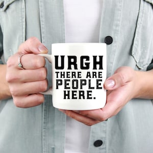 K&ouml;nnte beinhalten: Eine wei&szlig;e Keramik-Kaffeetasse mit schwarzem Text, der "URGH THERE ARE PEOPLE HERE." lautet.
