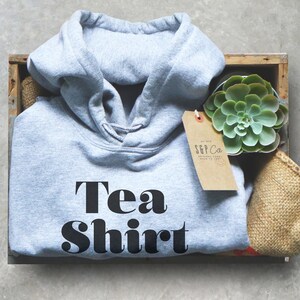 May include: A light grey hooded sweatshirt with the text "Tea Shirt" in black block letters. The sweatshirt is in a wooden crate with a small succulent plant and a brown paper tag.