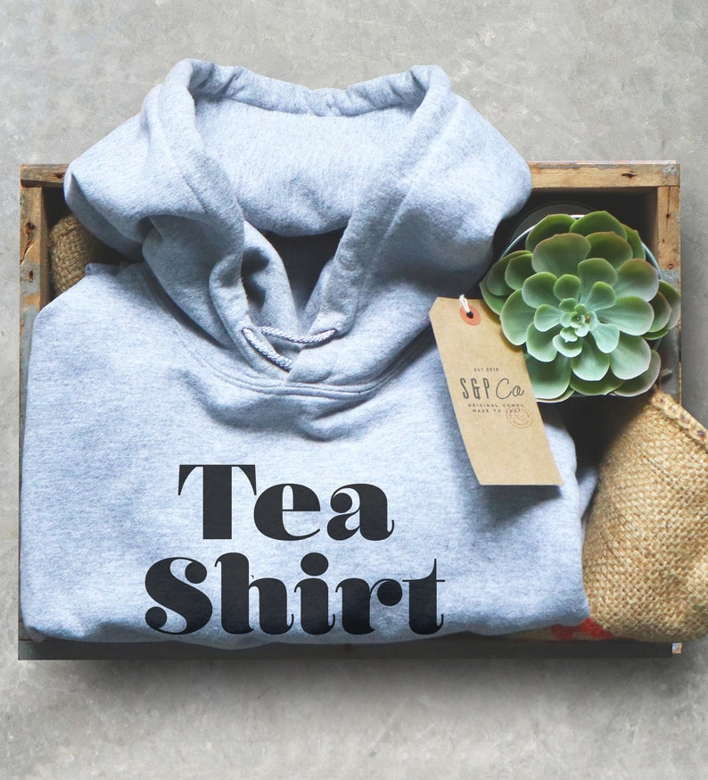 May include: A light grey hooded sweatshirt with the text "Tea Shirt" in black block letters. The sweatshirt is in a wooden crate with a small succulent plant and a brown paper tag.