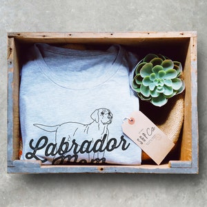 May include: A gray t-shirt with a black outline of a Labrador Retriever and the text "Labrador Mom". The t-shirt is in a wooden crate with a small succulent plant and a brown tag with the text "SFF Co."
