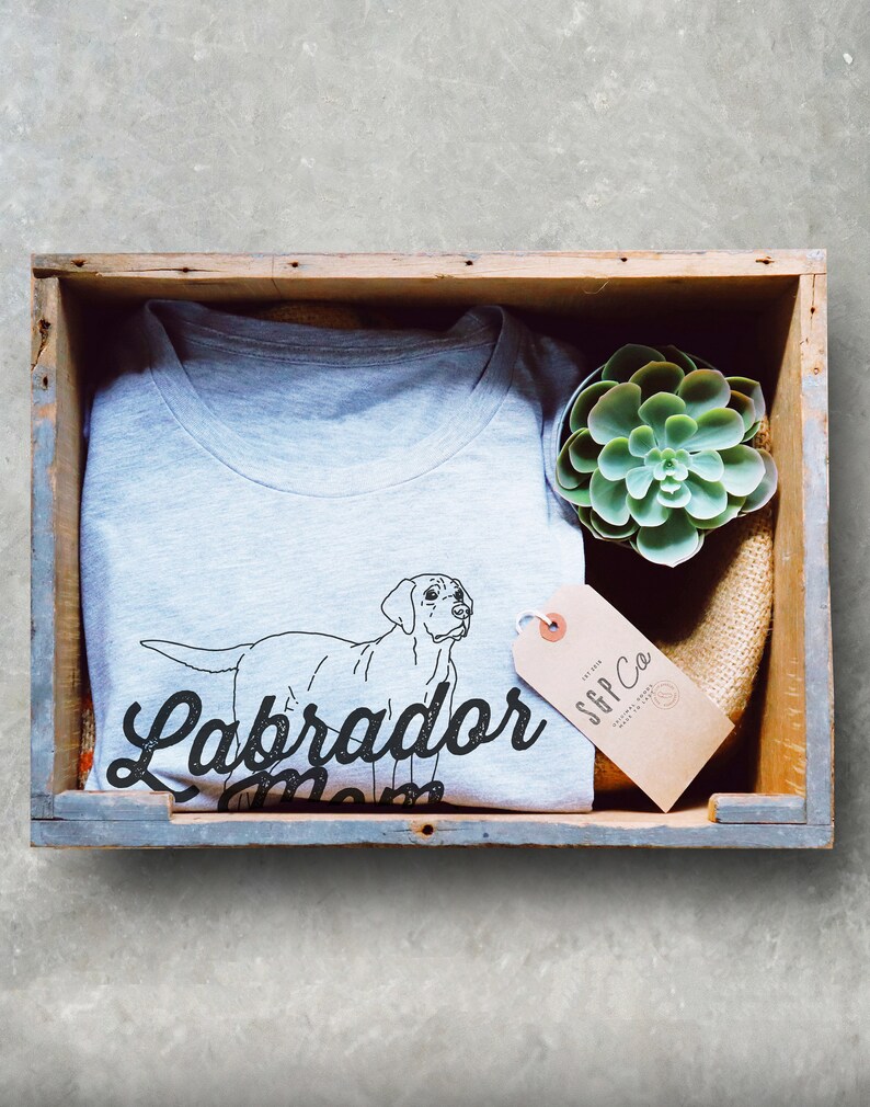 May include: A gray t-shirt with a black outline of a Labrador Retriever and the text "Labrador Mom". The t-shirt is in a wooden crate with a small succulent plant and a brown tag with the text "SFF Co."