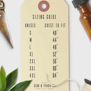 Op de afbeelding: Een beige papieren label met een maattabel voor unisex kleding. Het label geeft maten S tot en met 4XL met de bijbehorende borstmaten in inches. Het label is gemarkeerd met "Stag & Peach Co."