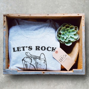May include: A gray t-shirt with the text "LET'S ROCK" and a drawing of a hand with bones. The t-shirt is in a wooden crate with a small succulent plant and a brown tag that says "SFF Co."