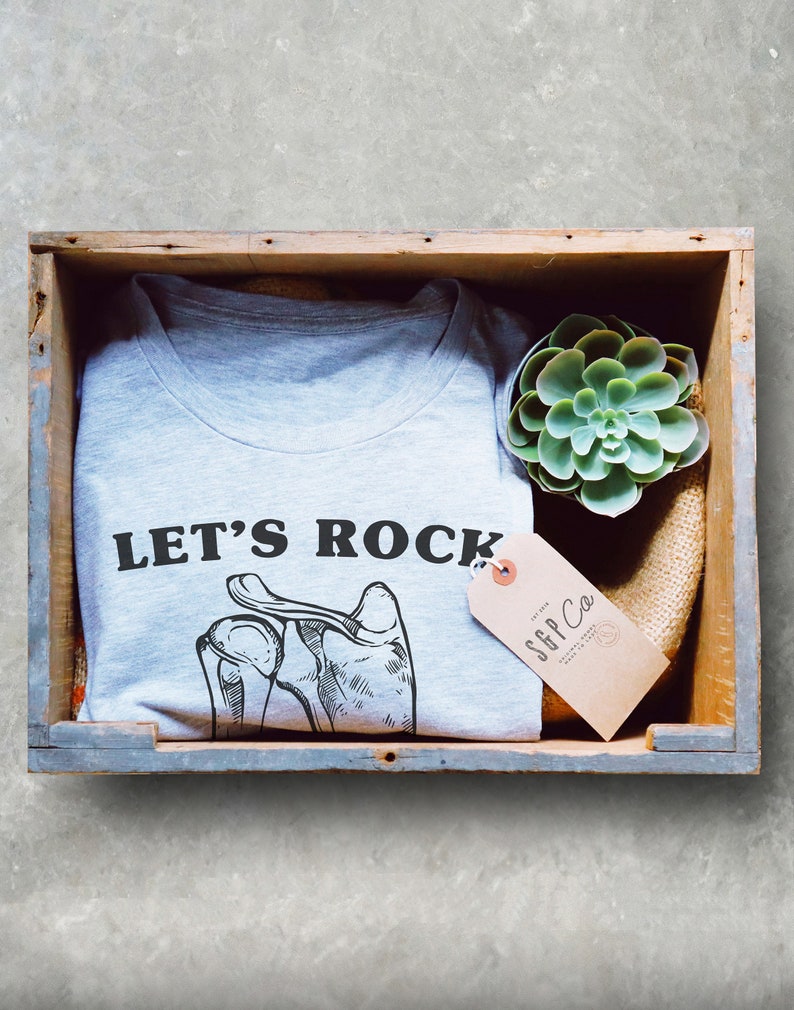 May include: A gray t-shirt with the text "LET'S ROCK" and a drawing of a hand with bones. The t-shirt is in a wooden crate with a small succulent plant and a brown tag that says "SFF Co."