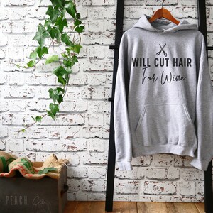 Op de afbeelding: Grijs hoodie met de tekst "WILL CUT HAIR for Wine" en een afbeelding van een schaar.