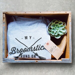 May include: A gray t-shirt with the text "My broomstick runs on" and a graphic of two crossed broomsticks. The t-shirt is in a wooden crate with a small succulent plant and a brown tag with the text "SGP Co."