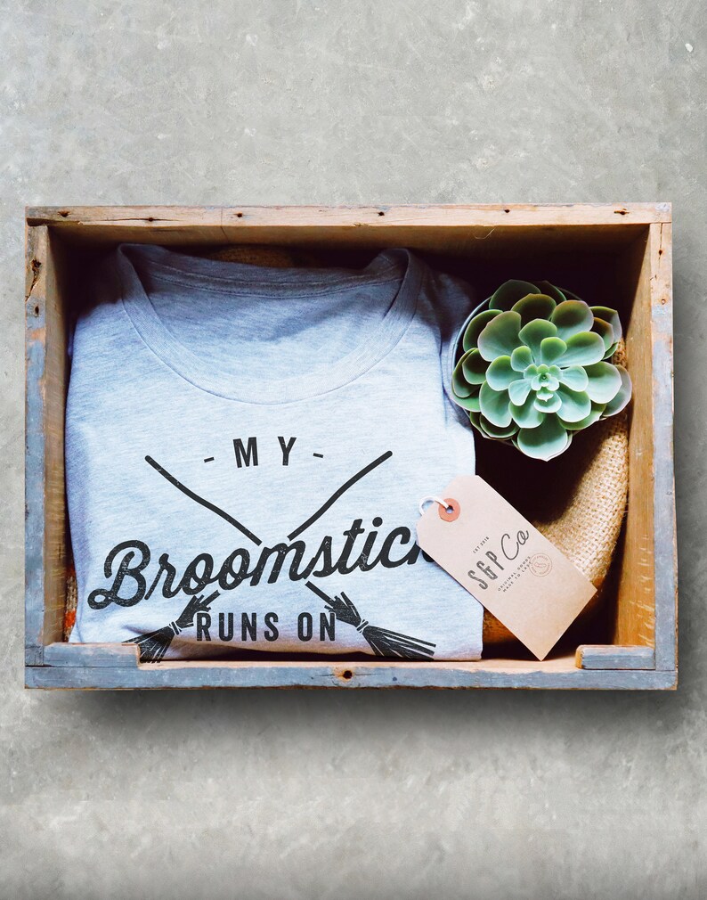 May include: A gray t-shirt with the text "My broomstick runs on" and a graphic of two crossed broomsticks. The t-shirt is in a wooden crate with a small succulent plant and a brown tag with the text "SGP Co."