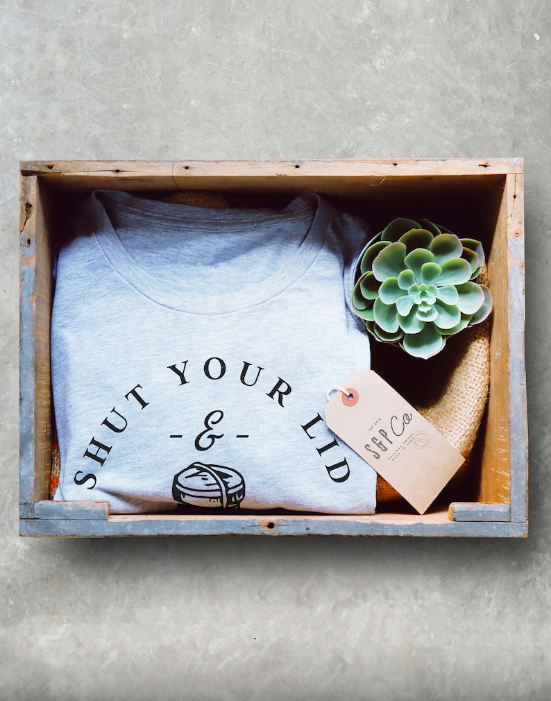 May include: A gray t-shirt with the text "Shut Your Lid - & -" and a small graphic of a lid. The t-shirt is in a wooden crate with a small succulent plant and a brown tag that says "S&P Co."