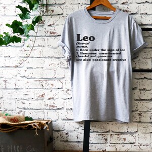 May include: Gray t-shirt with black text that reads "Leo (lee-o) noun. 1. Born under the sign of leo 2. Humorous, warm-hearted cheerful and generous see also: passionate creative"