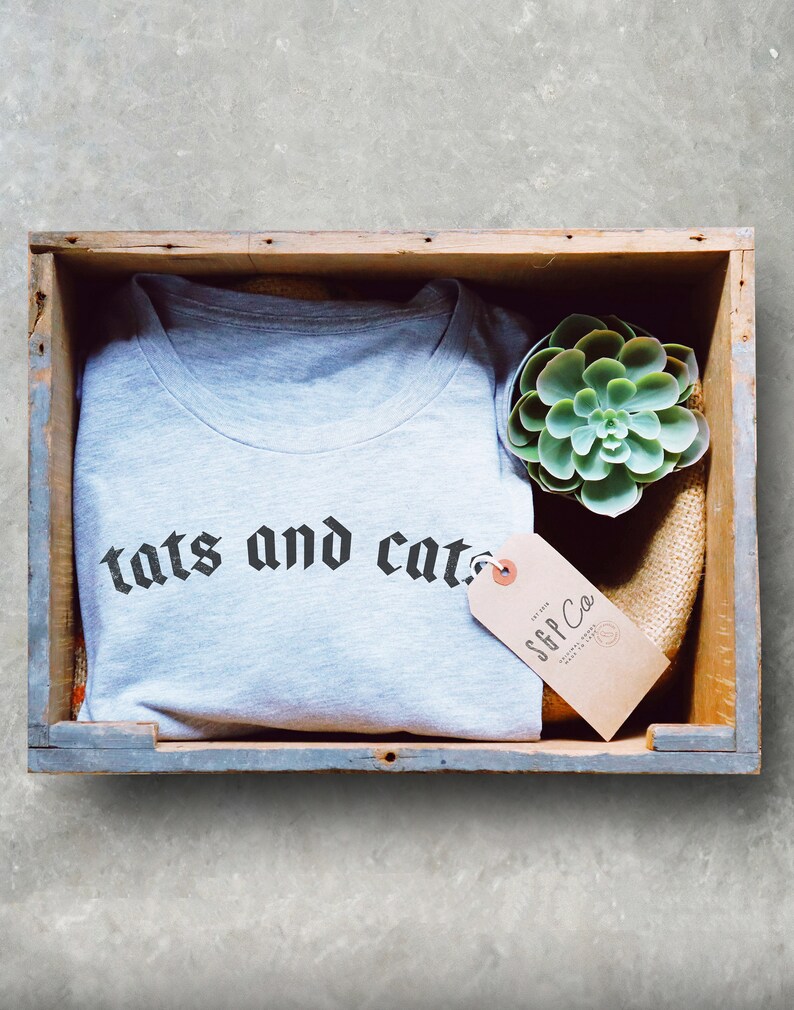 May include: A grey t-shirt with the text "tats and cats" in black gothic font. The shirt is folded and placed in a wooden crate with a small succulent plant and a brown tag that reads "SFFP Co."