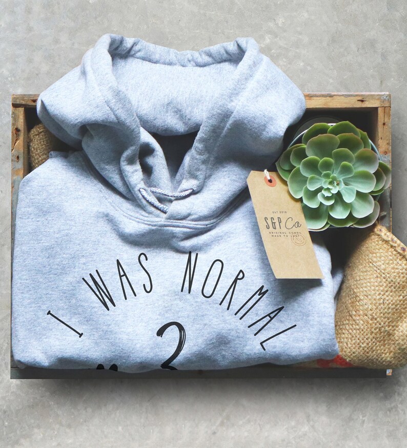 May include: A light gray hoodie with the text "I was normal 2" printed on the front. The hoodie is in a wooden box with a small succulent plant and a brown tag with the text "S&P Co."