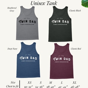 May include: A unisex tank top with the text "Twin Dad" and "Always Overachieving" in black lettering on a heather grey, classic black, deep navy, and classic red tank top. The tank tops are shown in various sizes from XS to XL with corresponding chest measurements in inches.