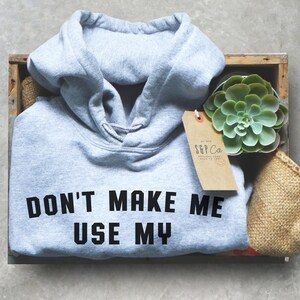 May include: A light gray hooded sweatshirt with the text "DON'T MAKE ME USE MY" printed in black on the front. The sweatshirt is folded and placed in a wooden crate with a small succulent plant and a brown paper tag.