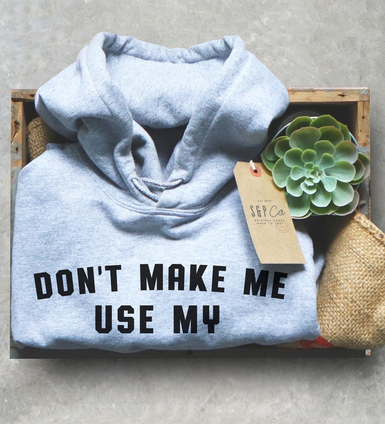 May include: A light gray hooded sweatshirt with the text "DON'T MAKE ME USE MY" printed in black on the front. The sweatshirt is folded and placed in a wooden crate with a small succulent plant and a brown paper tag.
