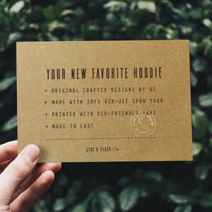 May include: A tan card with the text "YOUR NEW FAVORITE HOODIE" and additional text about the hoodie's features. The card is held in a hand against a blurred green background. The card also has the text "STAG & PEACH Co."