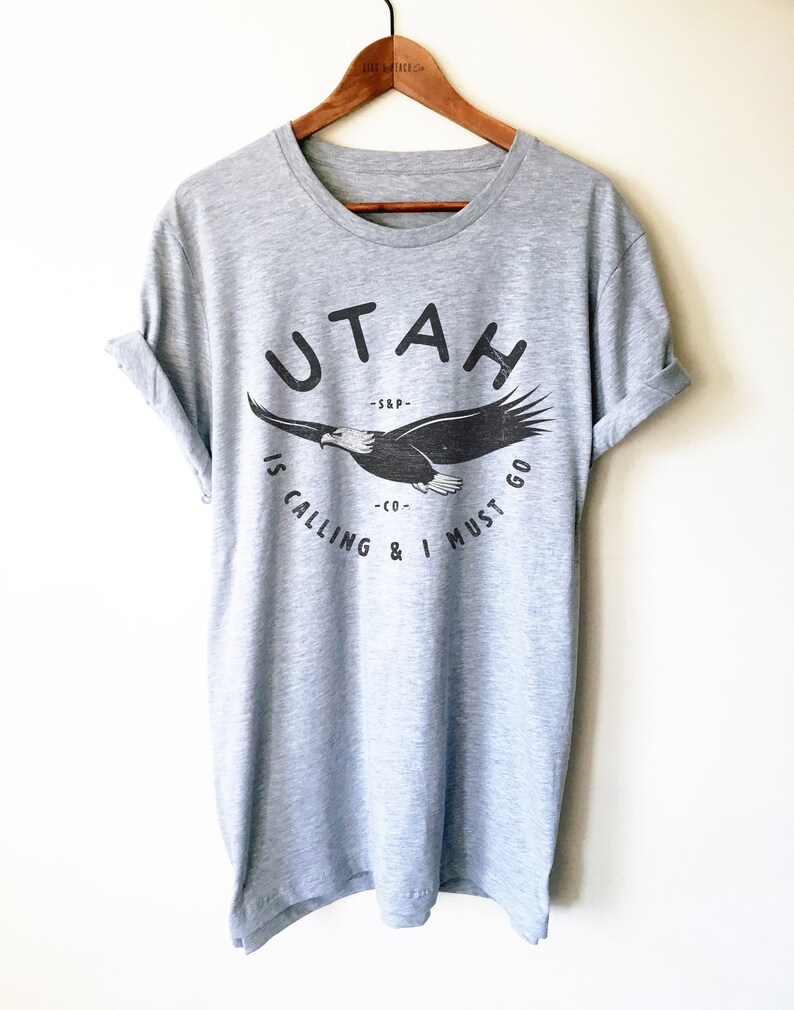 Op de afbeelding: Grijs T-shirt met een zwarte afbeelding van een vliegende adelaar met de tekst "UTAH IS CALLING & I MUST GO" gedrukt op de voorkant.
