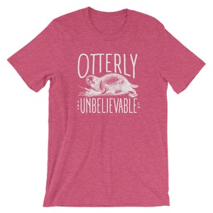 Puede incluir: Una camiseta rosa jaspeada con un gr&aacute;fico blanco de una nutria y el texto "OTTERLY UNBELIEVABLE CO."