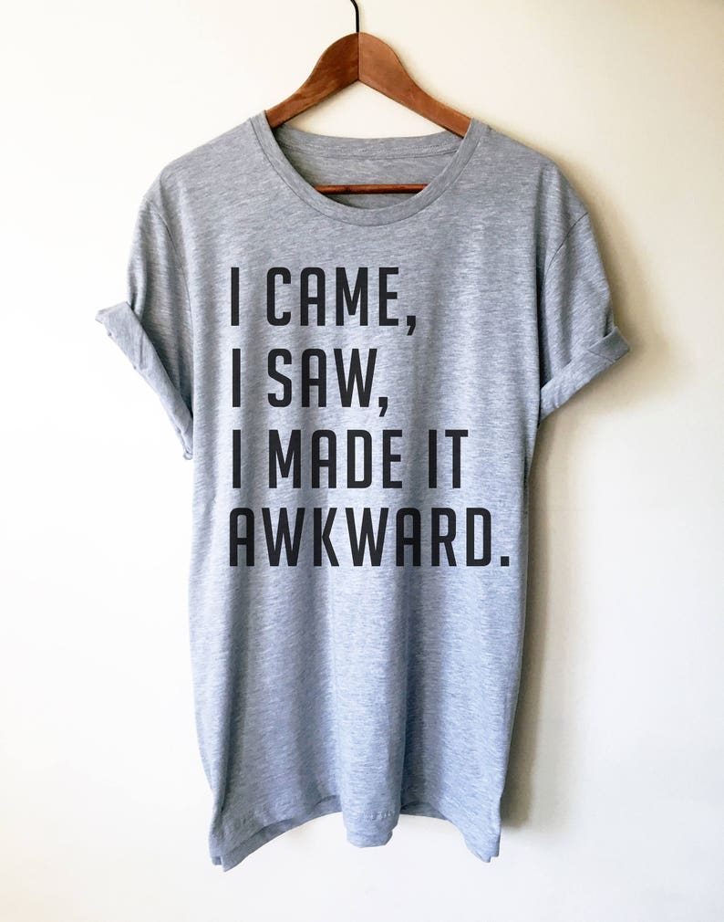 Puede incluir: Una camiseta gris jaspeada con el texto negro "I CAME, I SAW, I MADE IT AWKWARD", colgada de una percha de madera. La camiseta tiene las mangas dobladas y cuello redondo.