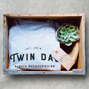 May include: A gray t-shirt with the text "Twin Da" and "Always Overachieving" in black lettering. The shirt is in a wooden crate with a small succulent plant and a brown tag that says "SGP Co."