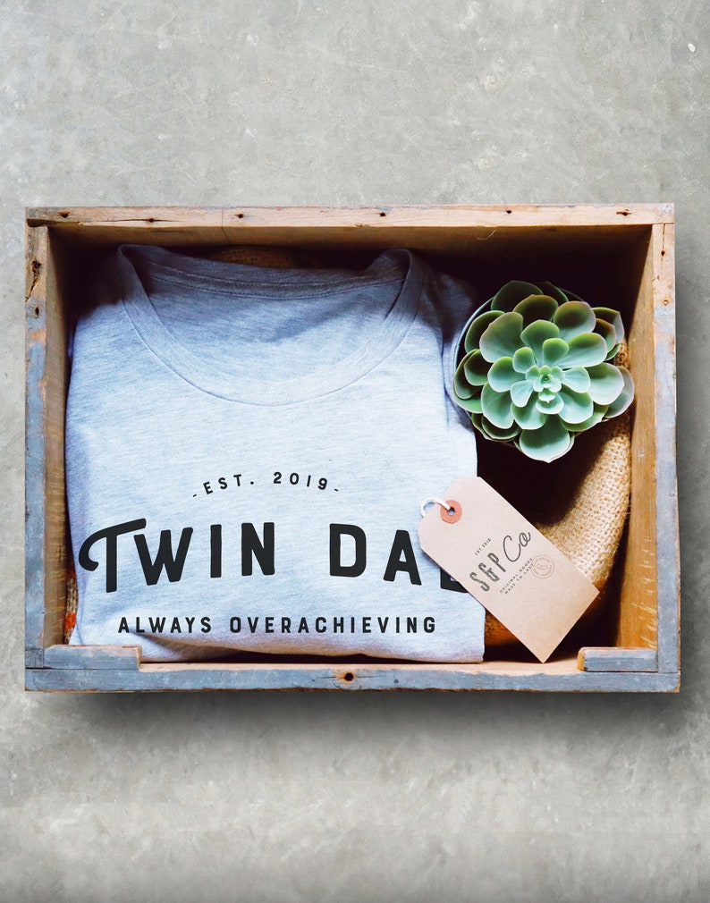 May include: A gray t-shirt with the text "Twin Da" and "Always Overachieving" in black lettering. The shirt is in a wooden crate with a small succulent plant and a brown tag that says "SGP Co."