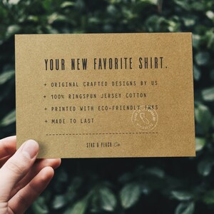 Może przedstawiać: Brązowa karta z tekstem "YOUR NEW FAVORITE SHIRT." i listą cech: "ORIGINAL CRAFTED DESIGNS BY US", "100% RINGSPUN JERSEY COTTON", "PRINTED WITH ECO-FRIENDLY INKS", "MADE TO LAST". Karta zawiera r&oacute;wnież tekst "STAG & PEACH Co."