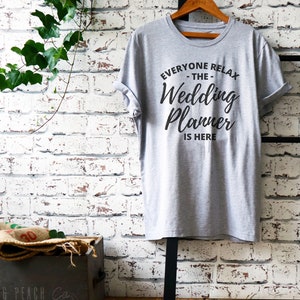 May include: A gray t-shirt with the text "Everyone Relax - The Wedding Planner Is Here" printed in black.