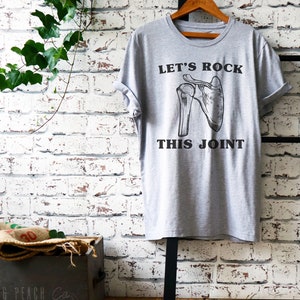 May include: Gray t-shirt with a black graphic of a shoulder joint and the text "Let's Rock This Joint".